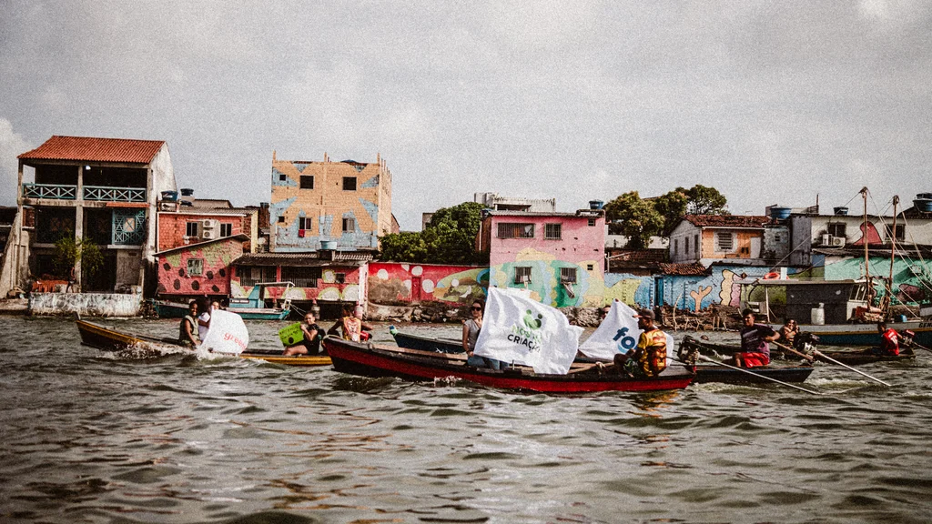 Barcos com bandeiras do Nós na Criação navegando diante de casas coloridas à beira d’água